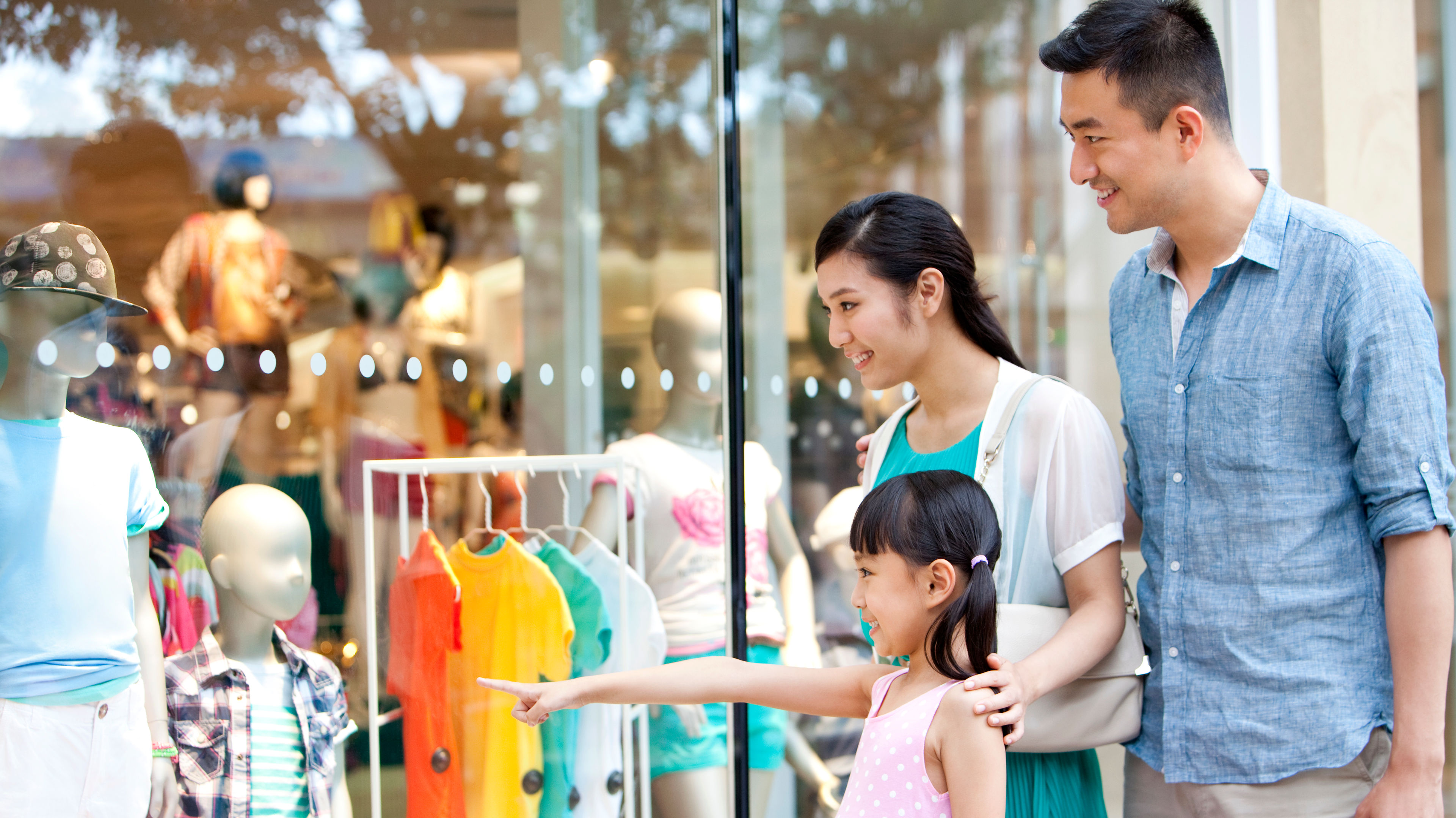 A smiling family with a young daughter points excitedly at a store window displaying colorful children's clothing